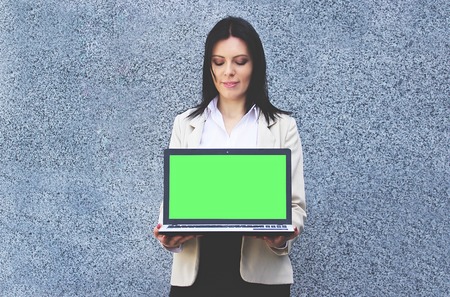 Technologies for business. Portrait of confident business woman in formal wear is keeping laptop with green chrome key screen to camera. Gray asphalt background.の写真素材