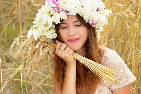 Enjoying the nature. Portrait of attractive young woman with flowers on her head keeping wheat in her hands while sitting in the yellow wheat field.の写真素材