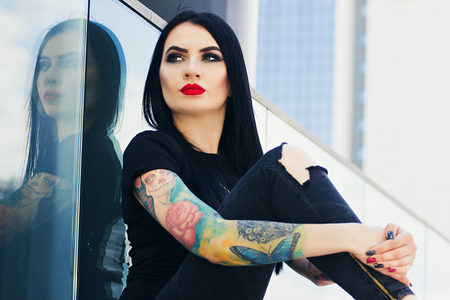 Tattoo fashion. Portrait of fashionable tattoed hipster girl with red lips sitting against urban background.の写真素材