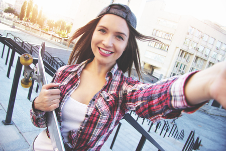 Happy moments must be saved. Cheerful, young and beautiful hipster woman in shirt and cap holding camera and taking selfie while keeping skateboard in another hand.の写真素材