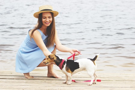 Let's play, honey. Horizontal shot of beautiful young woman in dress playing with dog and smiling while sitting on wooden pier.の写真素材