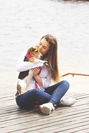 Hugging with pet. Full-length of attractive young woman in casual wear is hugging her little dog and smiling while sitting on wooden pier.の写真素材