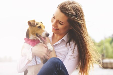 Me and my lovely pet. Portrait of young attractive woman in casual wear looking at dog and smiling while keeping dog on her hands, sitting on pier.の写真素材