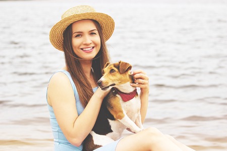 Me and my dog. Portrait of tender, young and beautiful woman in hat is keeping dog in hands and smiling while sitting near water on the beach.の写真素材