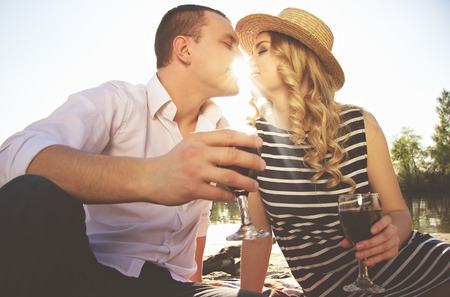 Happy couple. Low angle view of beautiful young woman and man keeping eyes closed and sitting close to each other while drinking wine on the beach. Evening sunlight between them.の写真素材
