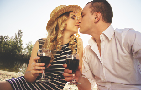 Love to kiss her lips. Portrait of man in casual wear kissing his young beautiful woman in dress and keeping glasses with wine in their hands while having picnic on beach.の写真素材