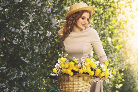 Perfect beauty. Portrait of attractive young woman in dress and hat keeping eyes closed and looking away while holding basket with flowers on bike.の写真素材