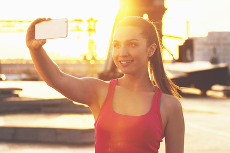 Sports selfie. Young and beautiful woman in sports wear looking at phone and smiling while taking selfie on the phone with evening sunlight on background.の写真素材