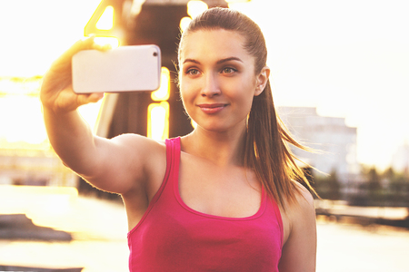 She loves selfie. Portrait of young and attractive woman in sports clothing looking at phone and smiling while taking selfie on her phone during outdoors workout.の写真素材