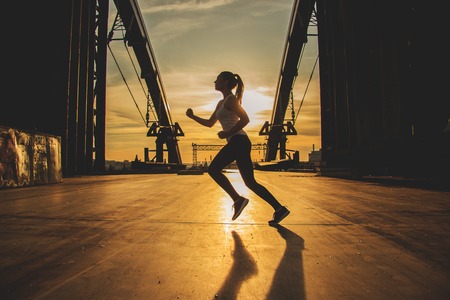 Evening jog. Side view of young attractive woman in sports clothing jogging on the bridge with industrial view and sunlight on background.の写真素材