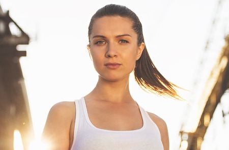 Confident sports woman. Portrait of young and attractive woman in sports clothing looking to camera while standing on bridge with sunlight on background.の写真素材