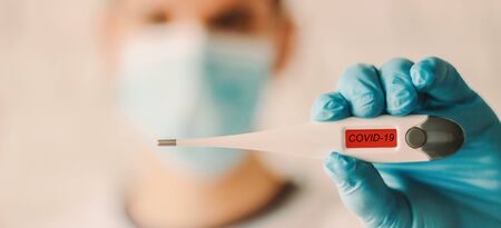 Closeup man in medical face mask and glove showing digital thermometer with COVID-19 text. Doctor in protective glove and mask holding electronic thermometer in hand. Coronavirus symptoms, diagnosisの写真素材