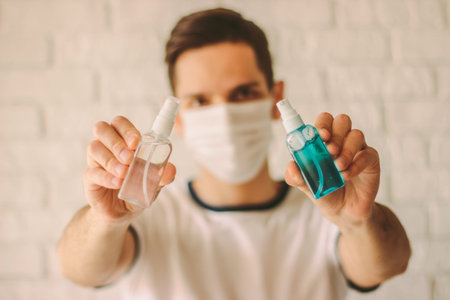 Young man in medical mask on face hold sanitizer bottles in hands. Professional doctor in protective...