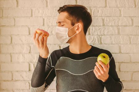 Man fitness trainer in medical face mask holds donut and apple in hands. Sports man in protective face mask choosing between healthy food or sugar snack. Dieting, home quarantine, coronavirus COVID-19の写真素材