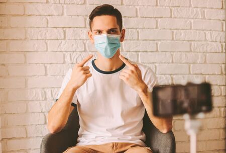Young man recording video instructions of prevention measures against coronavirus COVID-19 for his blog on his smart phone on tripod. Handsome internet blogger pointing on medical mask on face. Vlogの写真素材