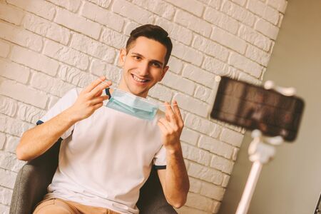 Handsome blogger wearing medical mask on face and recording video content on smart phone camera. Young man filming vlog with instructions of preventive measures against coronavirus COVID-19, nCov-19の写真素材