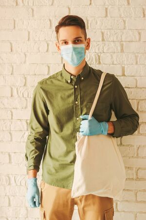 Young man in protective face mask and medical gloves carry cotton eco bag. Hipster man in medical mask on face and protective gloves hold tote bag. Personal protection, shopping, COVID-19, coronavirusの写真素材