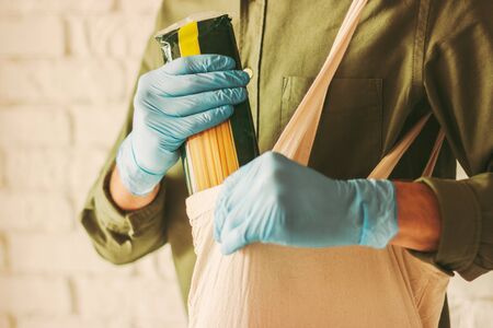 Closeup of young man shopper in medical protective gloves putting macaroni in cotton tote eco bag in grocery store. Hipster man shopping in food store. Personal safety, coronavirus COVID-19 protectionの写真素材