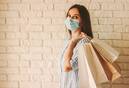 Beautiful girl in protective face mask holding shopping bags in hands. Young woman shopaholic in medical mask on face with paper bags. Female shopper. Safe shopping, coronavirus COVID-19, copy spaceの写真素材