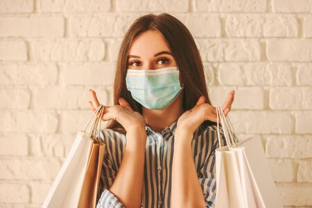 Beautiful girl shopper in medical face mask holding shopping bags in hands. Young happy woman customer in protective mask on face with paper bags. Girl shopaholic. Coronavirus COVID-19, sale, discountの写真素材
