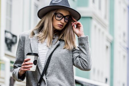 Tender and stylish girl. Portrait of young fashionable woman in coat and hat keeping eyes closed and holding paper cup of coffee in hand while standing outdoors. Hipster girl walking in the city.の写真素材