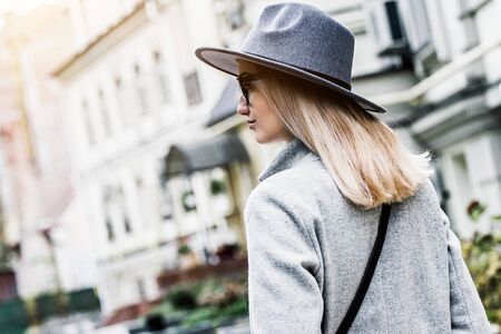 Back view of fashionable young woman in coat and hat looking away and walking on the city streets. Attractive and stylish hipster girl in modern urban outfit standing outdoors. City style and fashionの写真素材