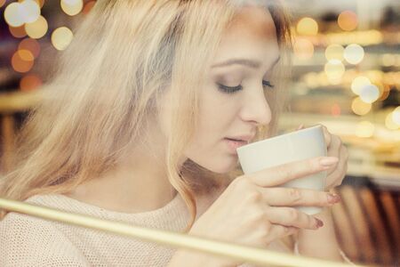 Beautiful blonde girl drinking coffee in cozy french cafe. Young woman enjoying tea in vintage european restaurant. Closeup of woman holding white cup in hands. Reflection view from outsideの写真素材