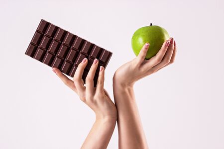 Deciding what to choose. Close up of sports woman hands with chocolate and apple isolated on white background. Hard choice between healthy and unhealthy food. Sweet temptation of fitness girlの写真素材