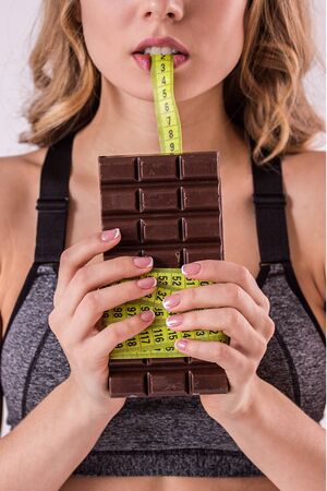 Craving to eat it. Young fitness woman holding chocolate bar with tape measure in hands isolated on white background. Hungry sports girl tempted by unhealthy sweet food. Dieting conceptの写真素材