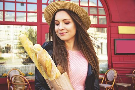 Absolute pure beauty. Closeup portrait of young and charming woman in pink dress and hat keeping baguettes in hands and keeping eyes closed while standing against French cafe.の写真素材