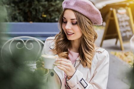 Enjoying nice day. Portrait of cute and beautiful woman in beret looking down and smiling while drinking coffee in french vintage cafe. Woman in french clothing. French fashion and style concept.の写真素材