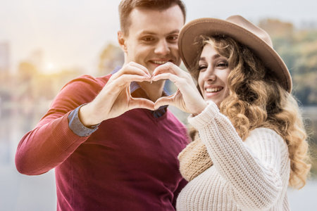 Showing symbol of love. Closeup young man and beautiful woman in hat keeping their hands together in...