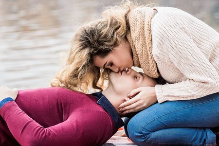 She loves to kiss his lips. Cropped image of beautiful blonde woman kissing her young boyfriend while sitting above him. Kissing couple in love. Togetherness concept. Romantic date outdoors.の写真素材