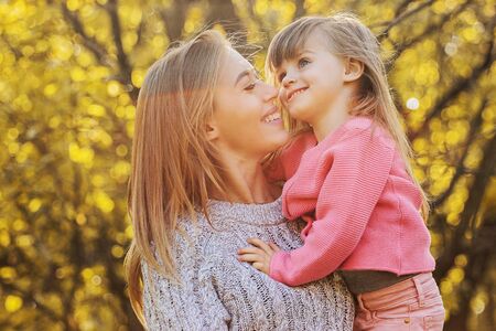 Portrait of cheerful young mother hugging her little daughter and holding her in hands while standing in autumn park. Mom and her kid having fun outdoors. Family relationships and love concept.の写真素材