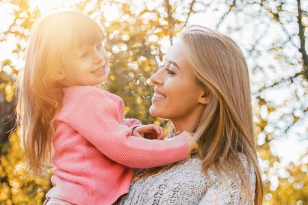 Happy young family embracing in autumn park. Happy young mother hugging her beautiful little daughter while standing outdoors. Cheerful woman holding her kid in hands. Mother love and happy familyの写真素材
