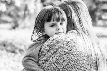 Black and white shot of young beautiful woman calming down and hugging her sad daughter while standing outdoors. Little disappointed girl embracing her lovely mom in autumn park. Family problemsの写真素材