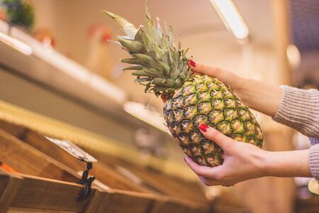 Closeup woman's hands holding pineapple with wooden market boxes on background. Young girl with exotic fruit in hands while doing shopping in supermarket. Healthy lifestyle and food, consumerism, saleの写真素材