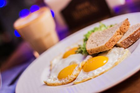 Closeup of fried eggs with toasted bread on white dish and glass with coffee on wooden table at modern restaurant. Traditional american breakfast in dark cafeの写真素材