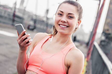 Portrait of happy young sports woman holding smart phone in hand and listening to music in earphones while standing on bridge. Attractive fitness girl smiles and listens motivational playlist on phoneの写真素材