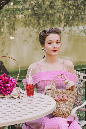 Portrait of stylish young tender woman with pin up hairstyle and pink vintage dress sitting at table in park and holding basket in hands. Beautiful romantic retro girl on picnic on summer gardenの写真素材