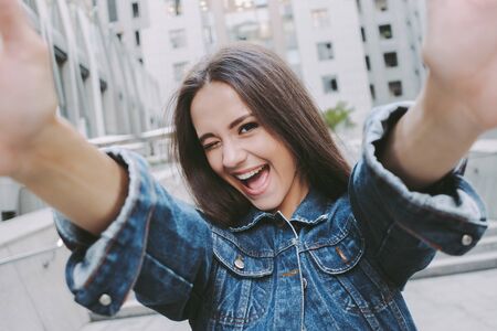 Young happy stylish woman winking and smiling while taking selfie on smart phone. Beautiful stylish cheerful hipster girl making portrait photo on camera. Having fun and making pictures on city streetの写真素材