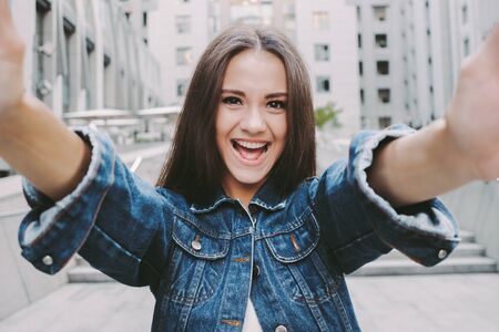 Cheerful young carefree woman making selfie on camera and smiling while having fun outdoors. Beautiful happy stylish hipster girl taking portrait photo on smart phone on city street. Positive girlの写真素材