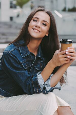 Happy young beautiful woman holding coffee cup in hands and smiling while sitting on city street. Attractive cheerful trendy hipster girl in stylish denim jacket drinks takeaway coffee or tea outdoorsの写真素材