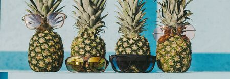 Closeup of group of fresh pineapple hipster family in funny and stylish sunglasses on table against blue bright wooden background. Funky, ripe, fashionable exotic fruits ananas. Summer island vacationの写真素材