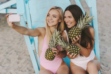 Two attractive women friends taking selfie together with pineapples in hand and smiling, sitting on lifeguard tower. Beautiful cheerful girls make sunny photos on smart phone. Summer vibes on beachの写真素材