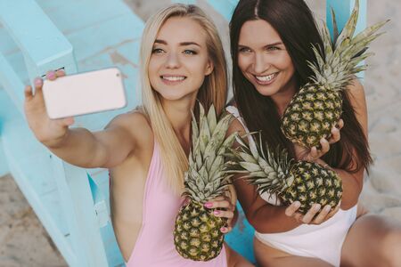 Happy young girls in swimsuits making sunny photos on smart phone camera at seaside. Beautiful cheerful women friends posing with pineapples in hands and taking selfie together on summer beachの写真素材