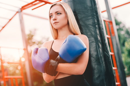 Portrait of confident young blonde fitness woman in boxing gloves standing on sports ground....