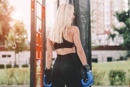 Back view of young beautiful sports woman having rest between boxing exercises on puch bag outdoors. Attractive blonde fitness girl in stylish clothing posing on sports ground. Sexy female boxerの写真素材