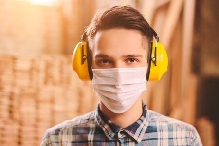 Portrait of confident man carpenter in medical face mask and hearing protectors at wooden workshop. Professional craftsman in protective mask and headphones at sawmill. Young male artisan at workplaceの写真素材