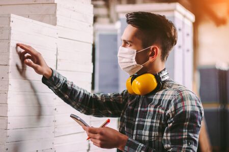 Portrait male foreman inspector in medical face mask and protective headphones checking woodwork stock at factory storage. Man supervisor counting wood inventory. Warehouse worker. COVID-19 quarantineの写真素材